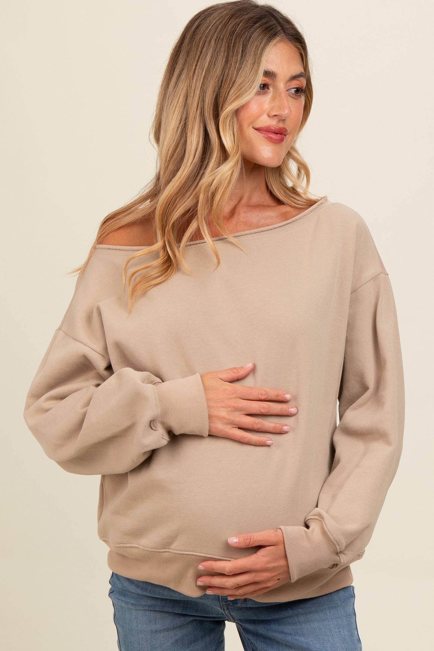 woman wearing a beige off-shoulder maternity sweatshirt and holding her baby bump