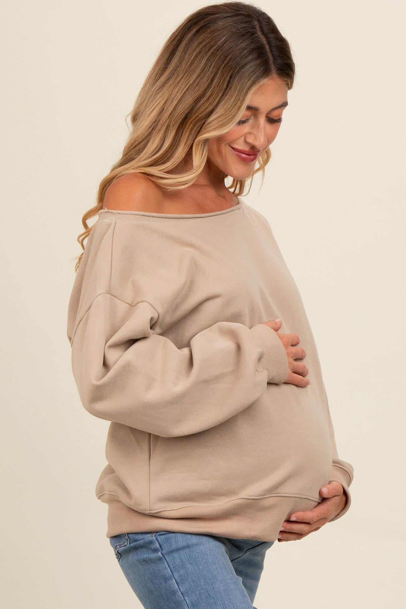 woman wearing a beige off-shoulder maternity sweater and holding her baby bump