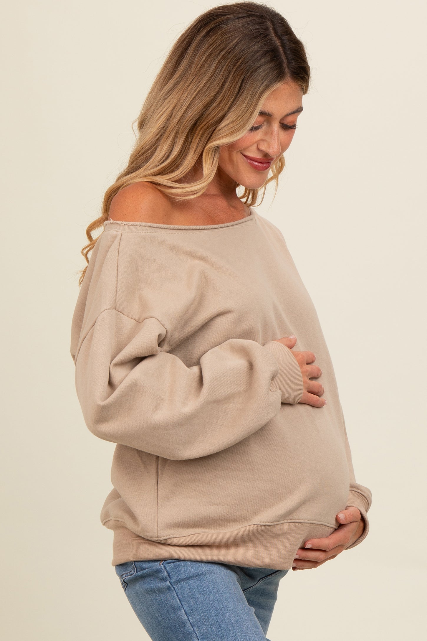 woman wearing a beige off-shoulder maternity sweater and holding her baby bump