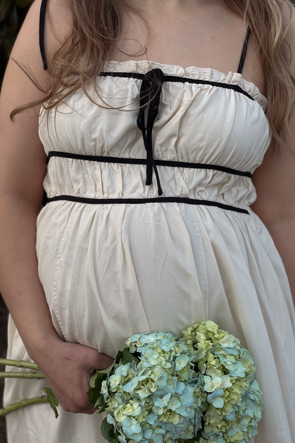 woman wearing a cream maternity dress with black accents and holding a bouquet of blue hydrangeas, showcasing her baby bump