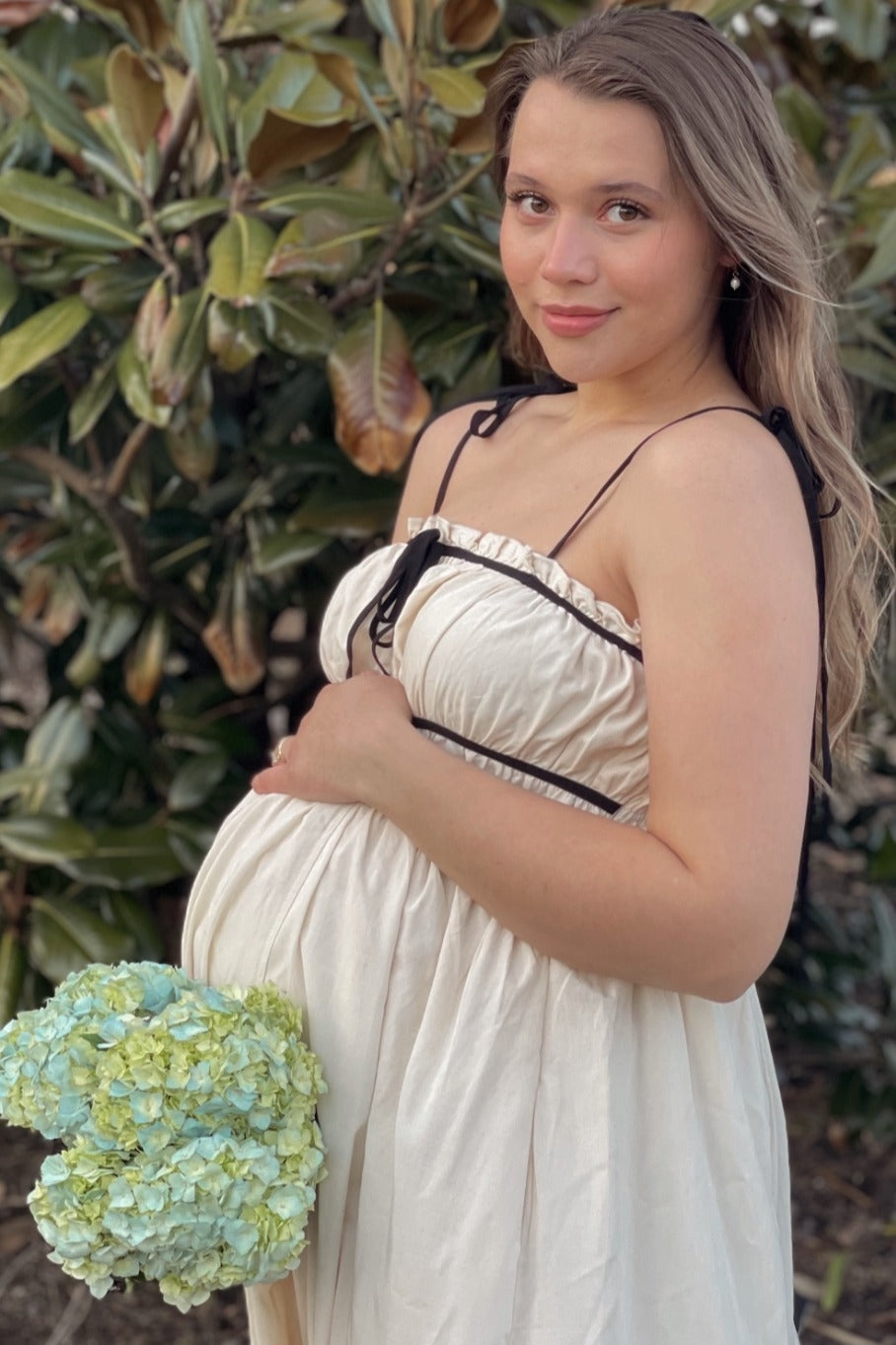 woman wearing a cream maternity dress with black accents, holding a bouquet of blue and green flowers and cradling her baby bump