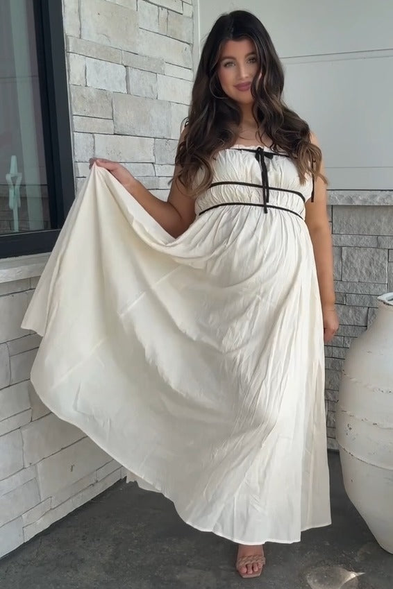woman wearing a cream maternity maxi dress with a ribbon tie, displaying her baby bump while standing outside