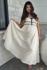 woman wearing a cream maternity maxi dress with a ribbon tie, displaying her baby bump while standing outside