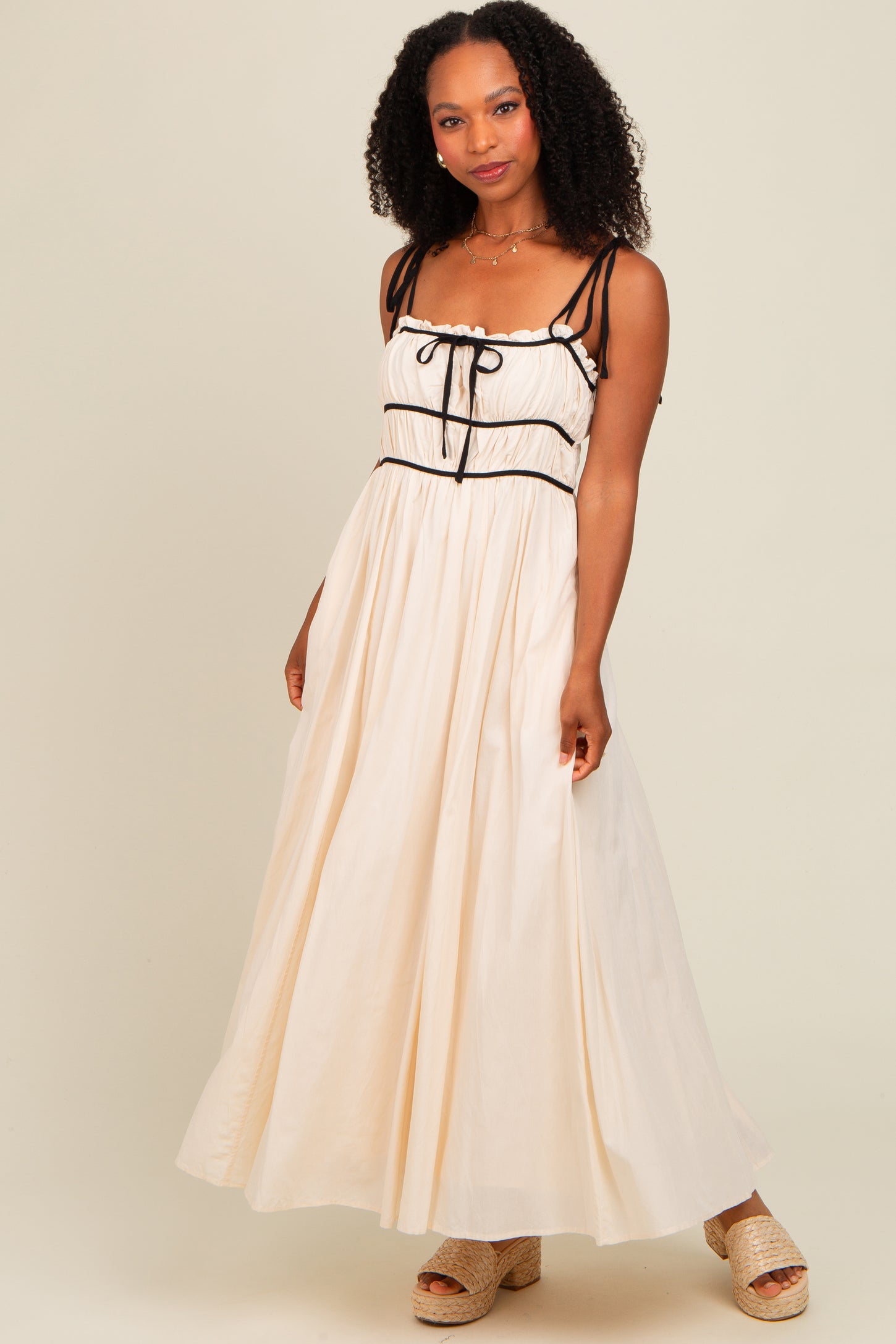 woman wearing a cream maxi dress with black accents and straps, standing on a neutral background