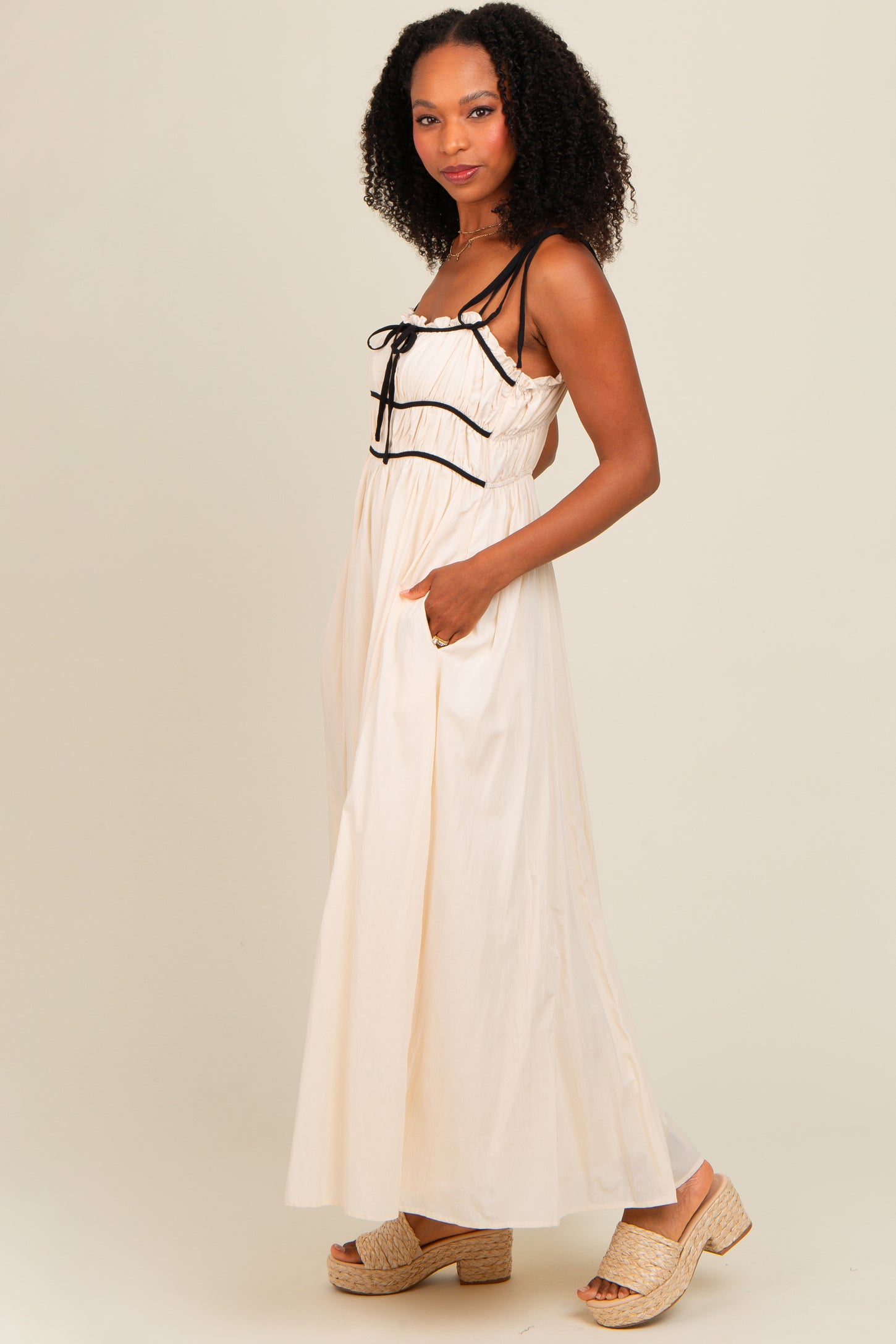 woman wearing a cream maxi dress with black drawstring details and pockets, paired with tan wedge sandals