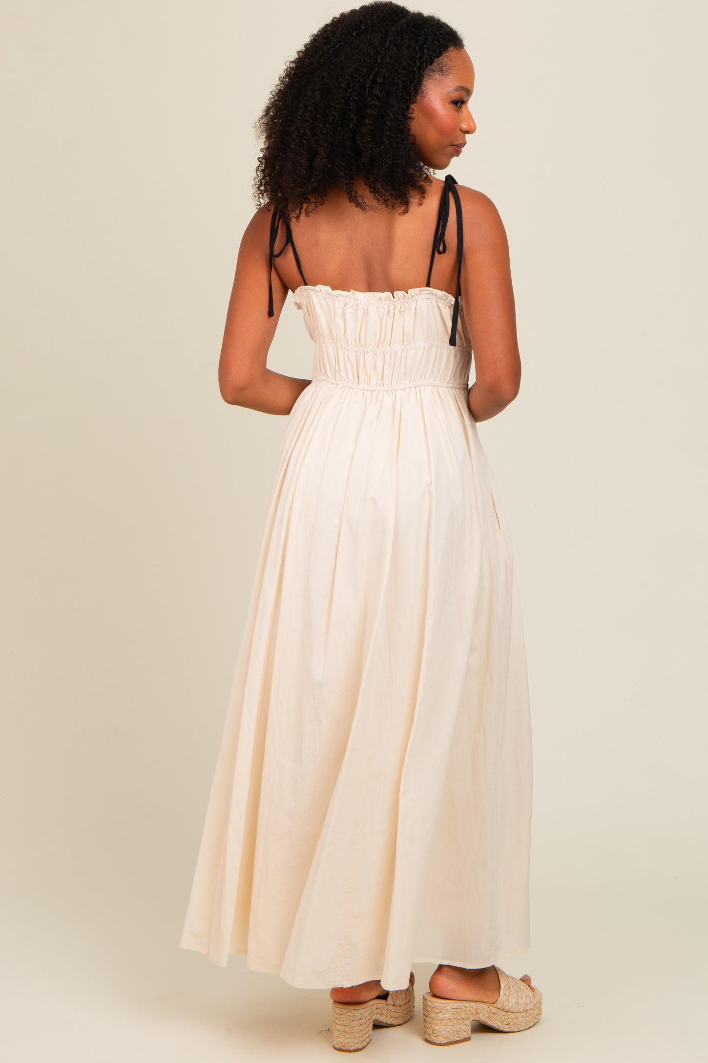 woman wearing a cream sleeveless maxi dress with shirred back and black straps, standing with her back to the camera