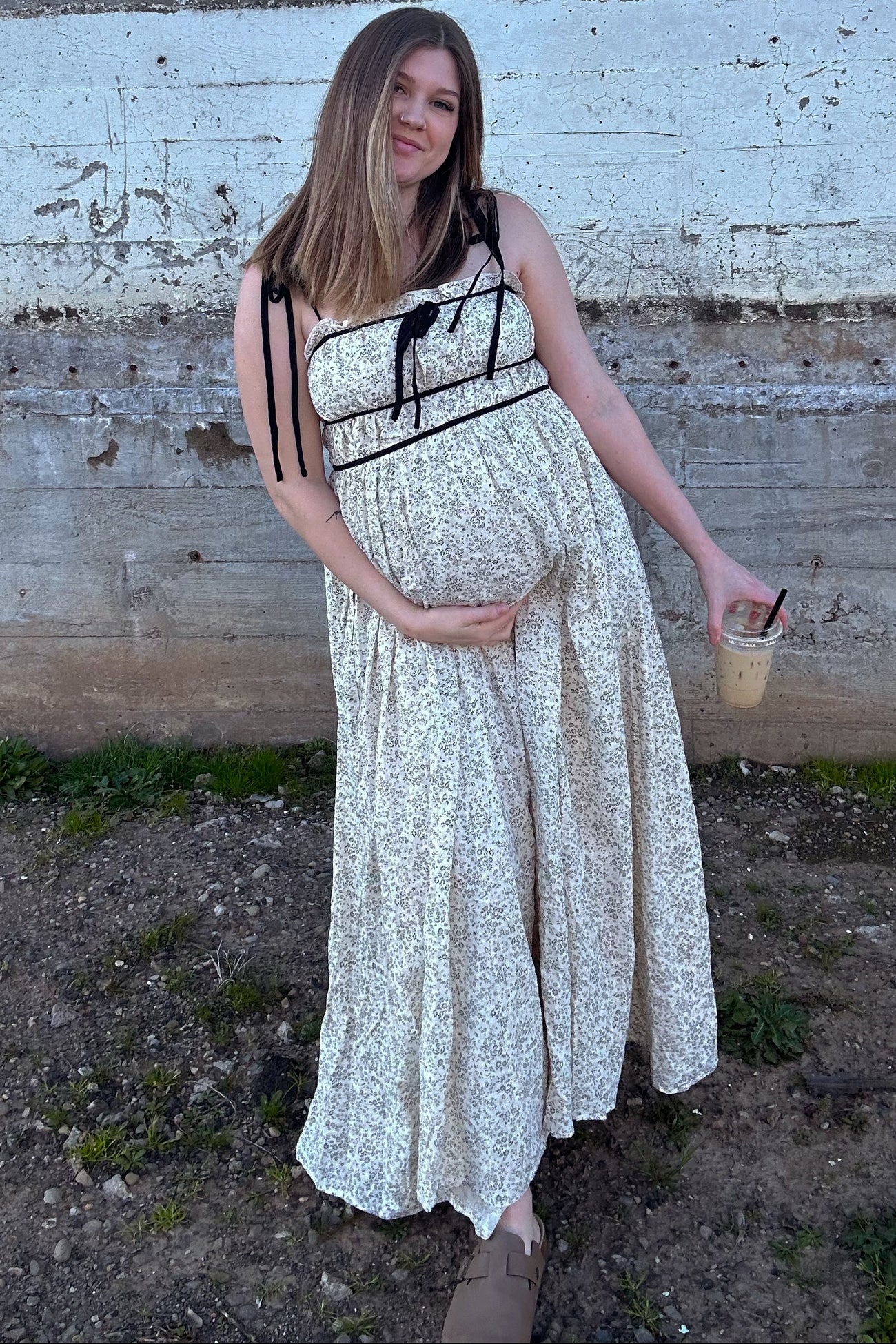 woman wearing a floral print maternity maxi dress and holding her baby bump while holding an iced drink