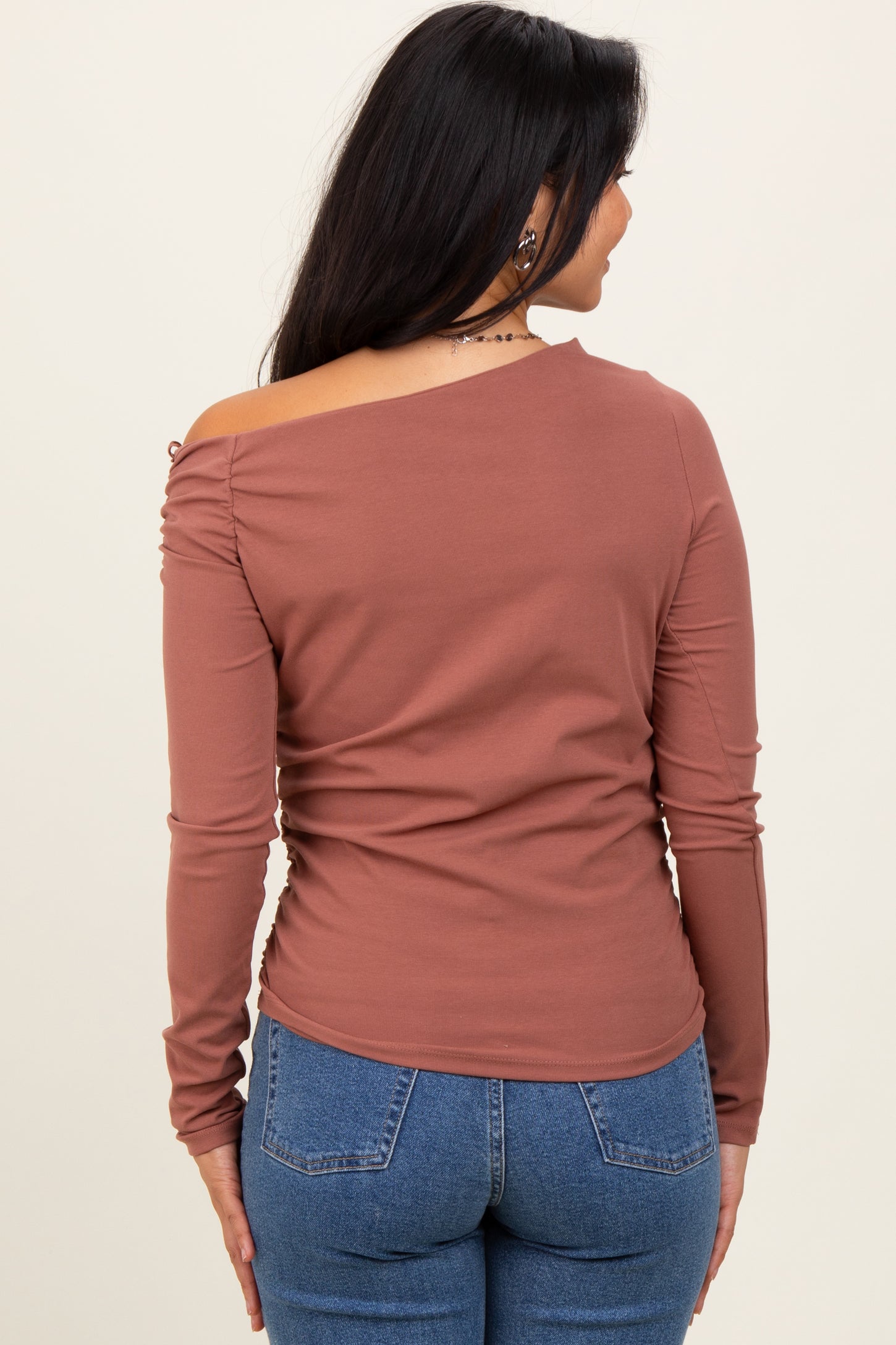 woman wearing a rust-colored long sleeve off-the-shoulder top with ruched details, paired with blue jeans