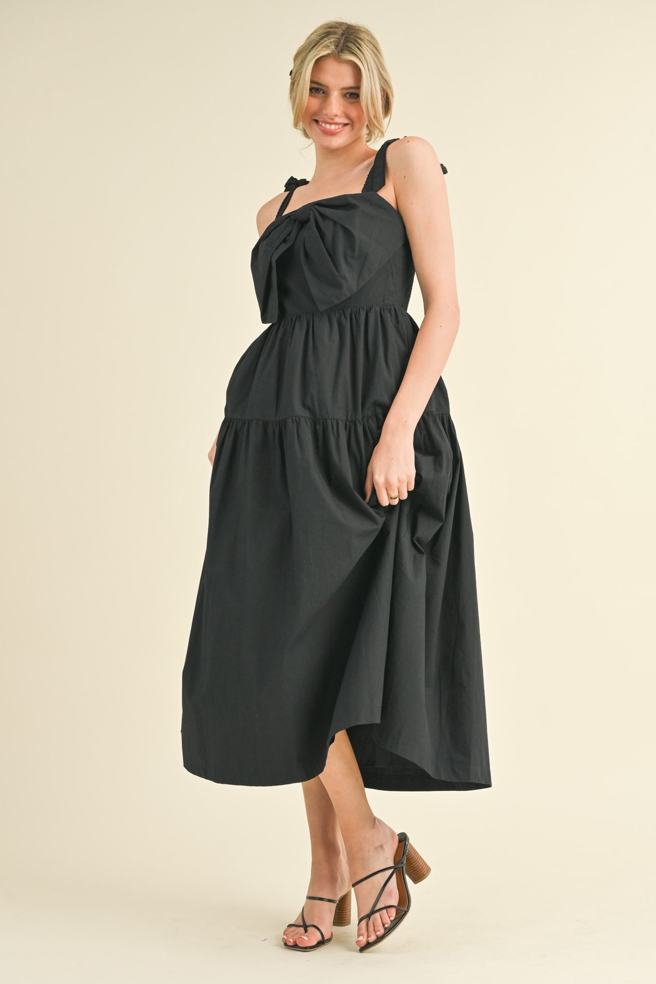 woman wearing a black tiered midi dress with bow straps and a flowing silhouette, standing on a light background