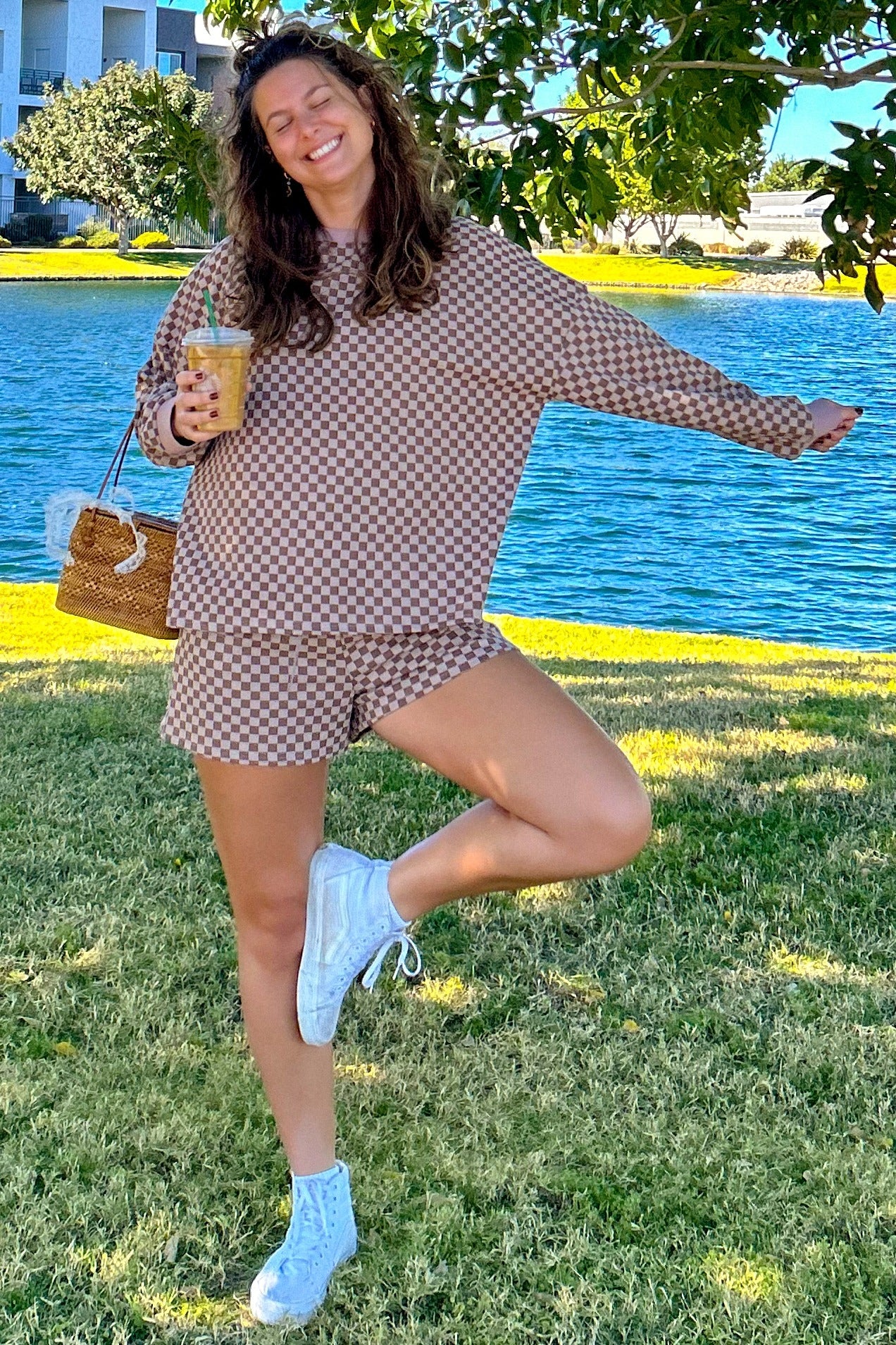 woman wearing a brown checkered oversized sweatshirt and matching shorts, holding a drink and posing by a body of water