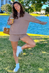 woman wearing a brown checkered oversized sweatshirt and matching shorts, holding a drink and posing by a body of water
