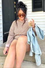 woman sitting on steps wearing a brown checkered long sleeve top and matching shorts, holding a drink and a denim jacket