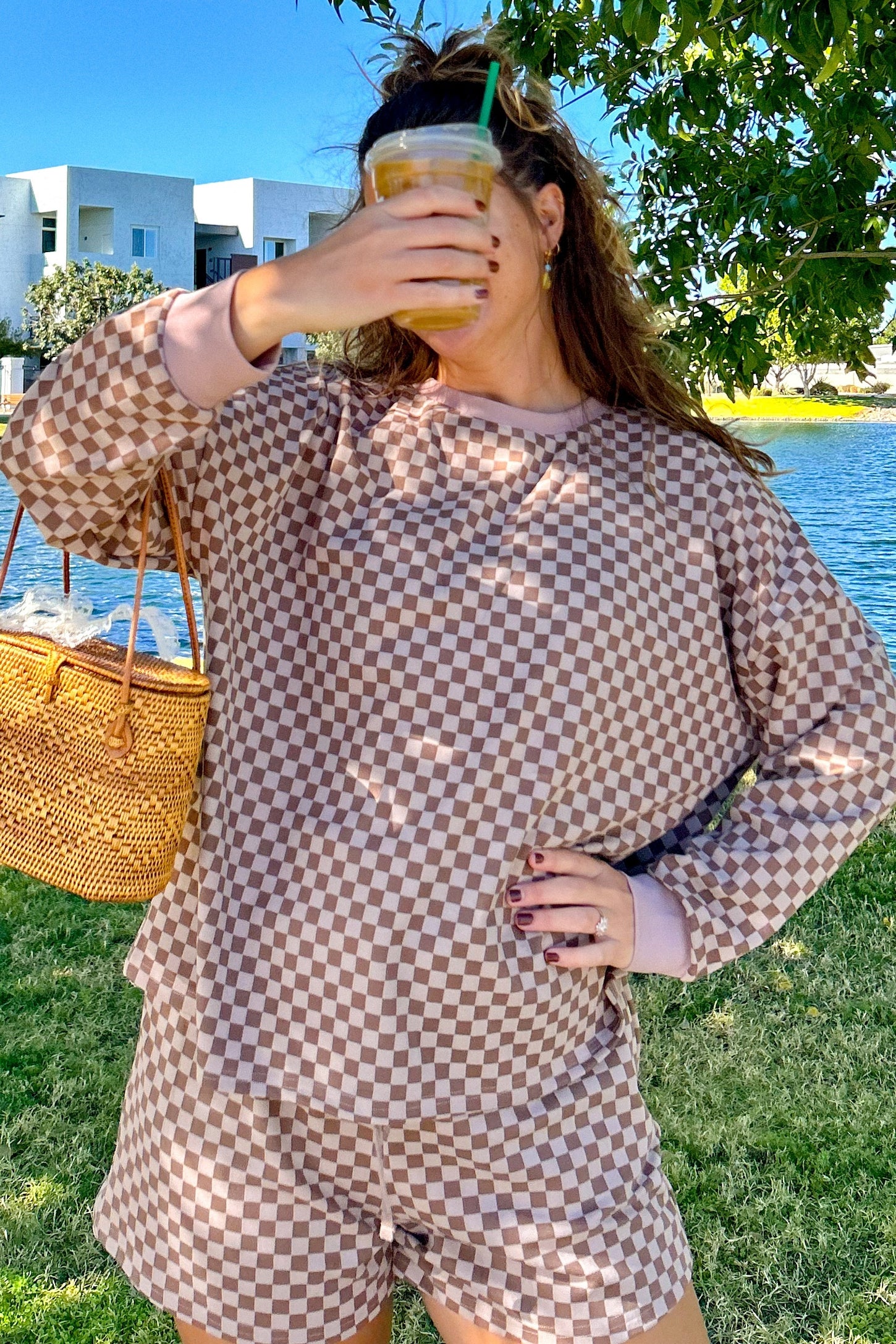 woman wearing a brown and cream checkered lounge set with long sleeves and shorts, holding a drink and a straw bag