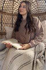 woman wearing a brown checkered sweatshirt seated in a cozy chair, holding a book and wrapped in a fluffy blanket