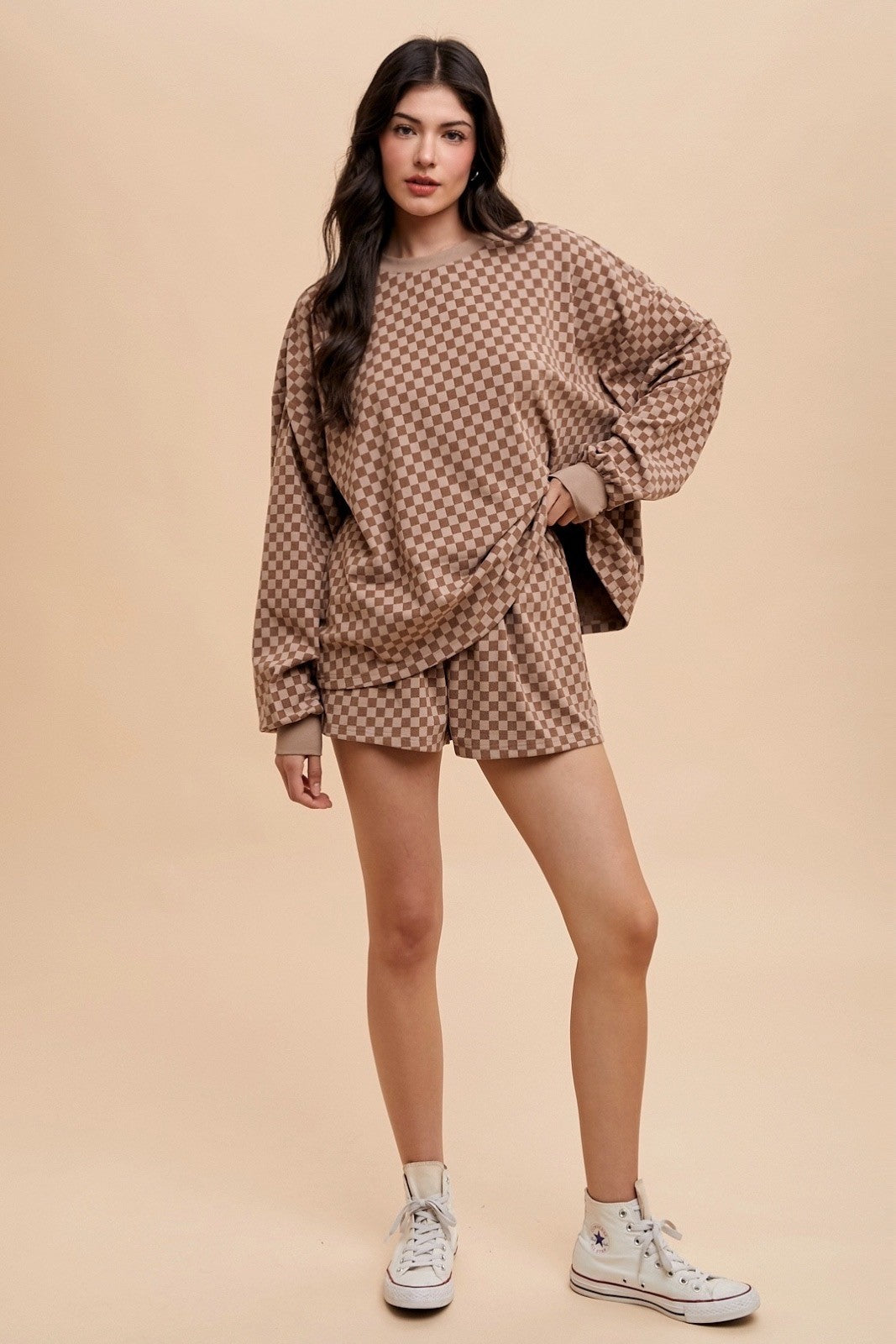 woman wearing a brown checkered oversized sweatshirt and matching shorts, standing against a light beige background