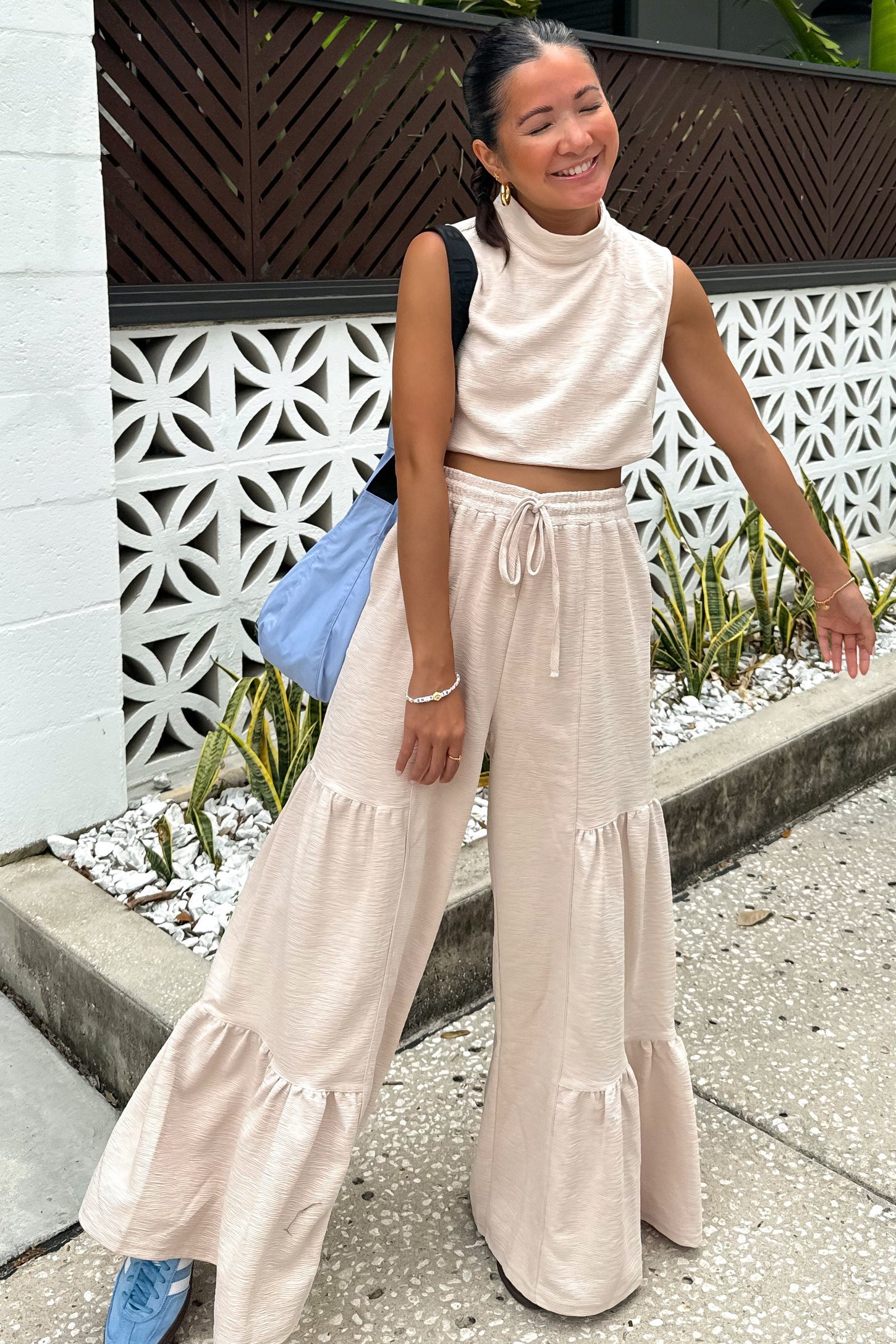 woman wearing a beige cropped sleeveless top and matching wide-leg pants with ruffle details, smiling outdoors