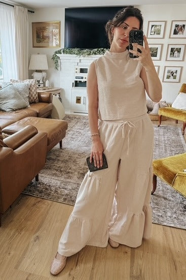 woman wearing a beige sleeveless top and matching wide-leg pants with ruffled hem, holding a clutch and posing in a living room