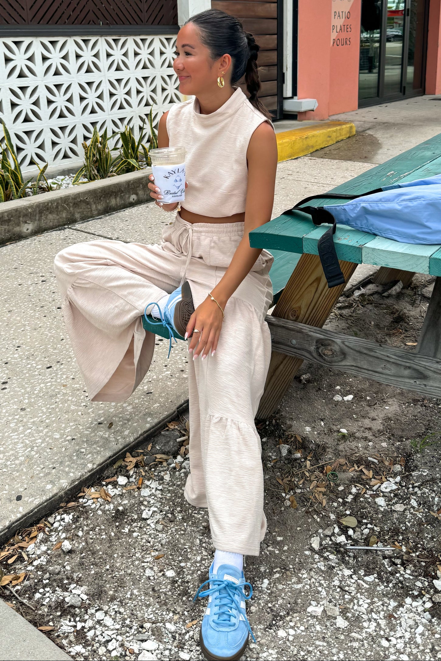 woman wearing a beige cropped sleeveless top and matching wide-leg pants, sitting on a bench with a drink in hand