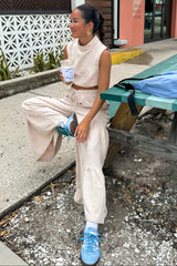 woman wearing a beige cropped sleeveless top and matching wide-leg pants, sitting on a bench with a drink in hand