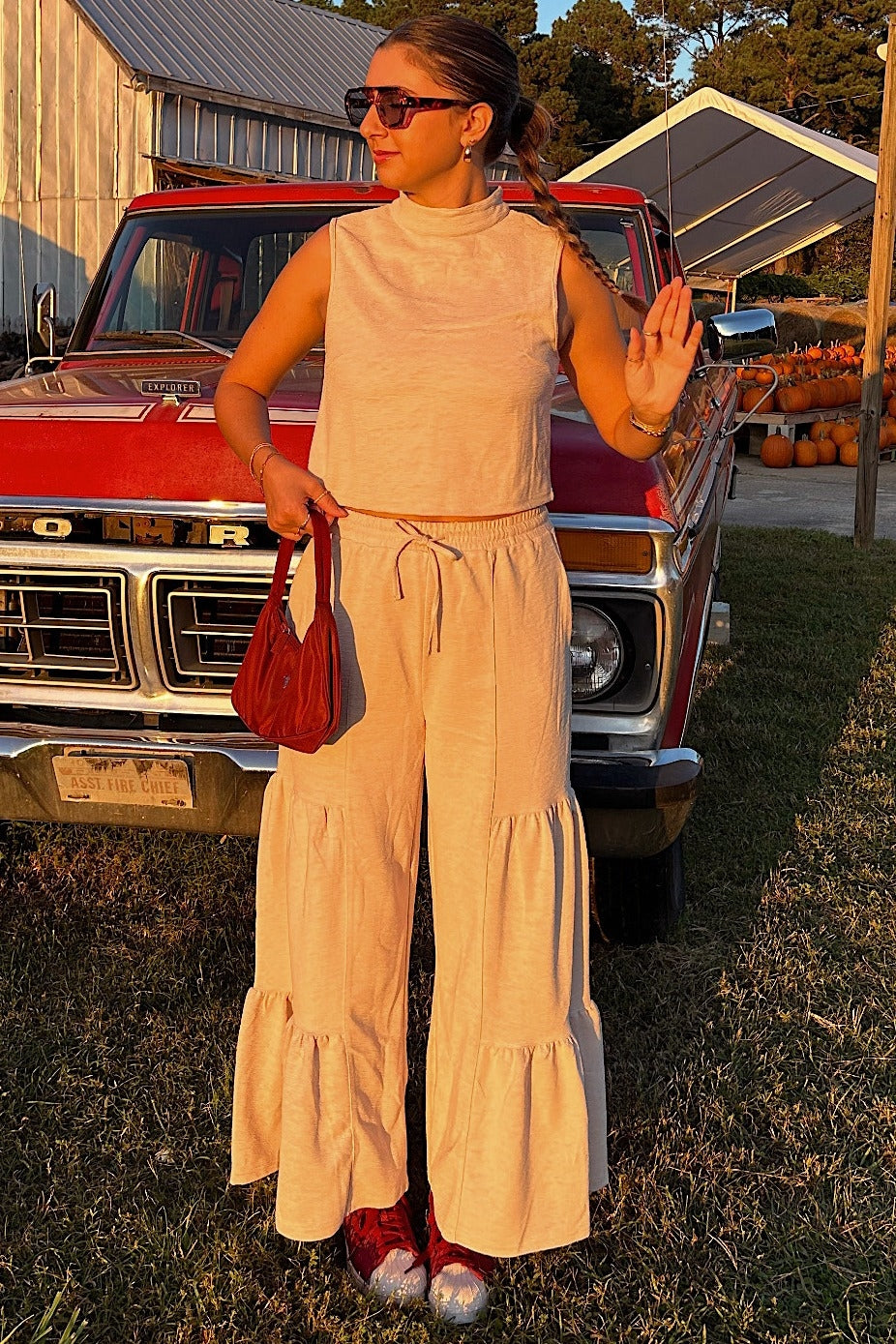 woman wearing a light beige sleeveless top and matching tiered wide-leg pants, holding a red bag and standing in a field