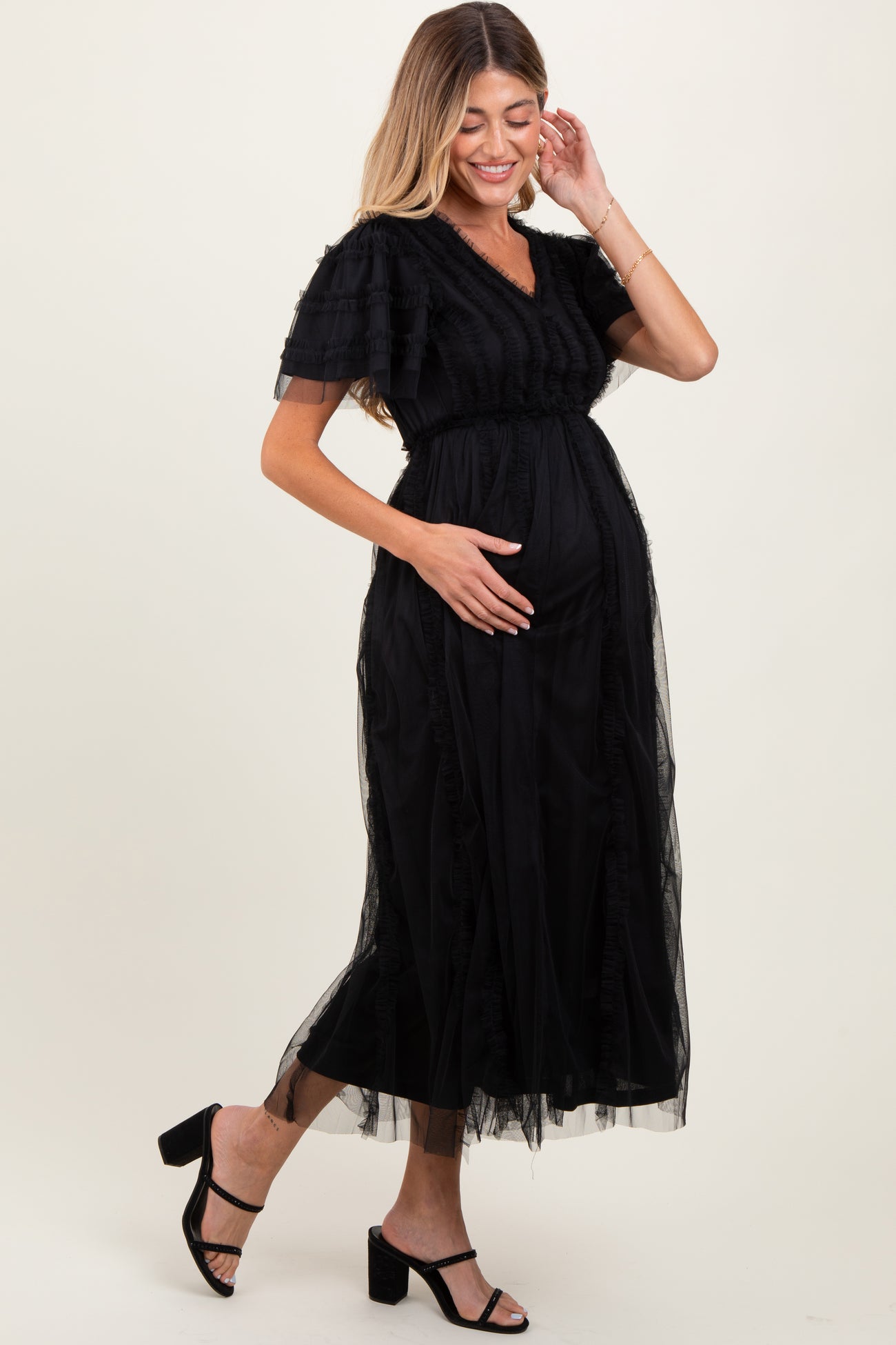 woman wearing a black maternity dress with tiered tulle details and a visible baby bump, posing with one hand on her belly