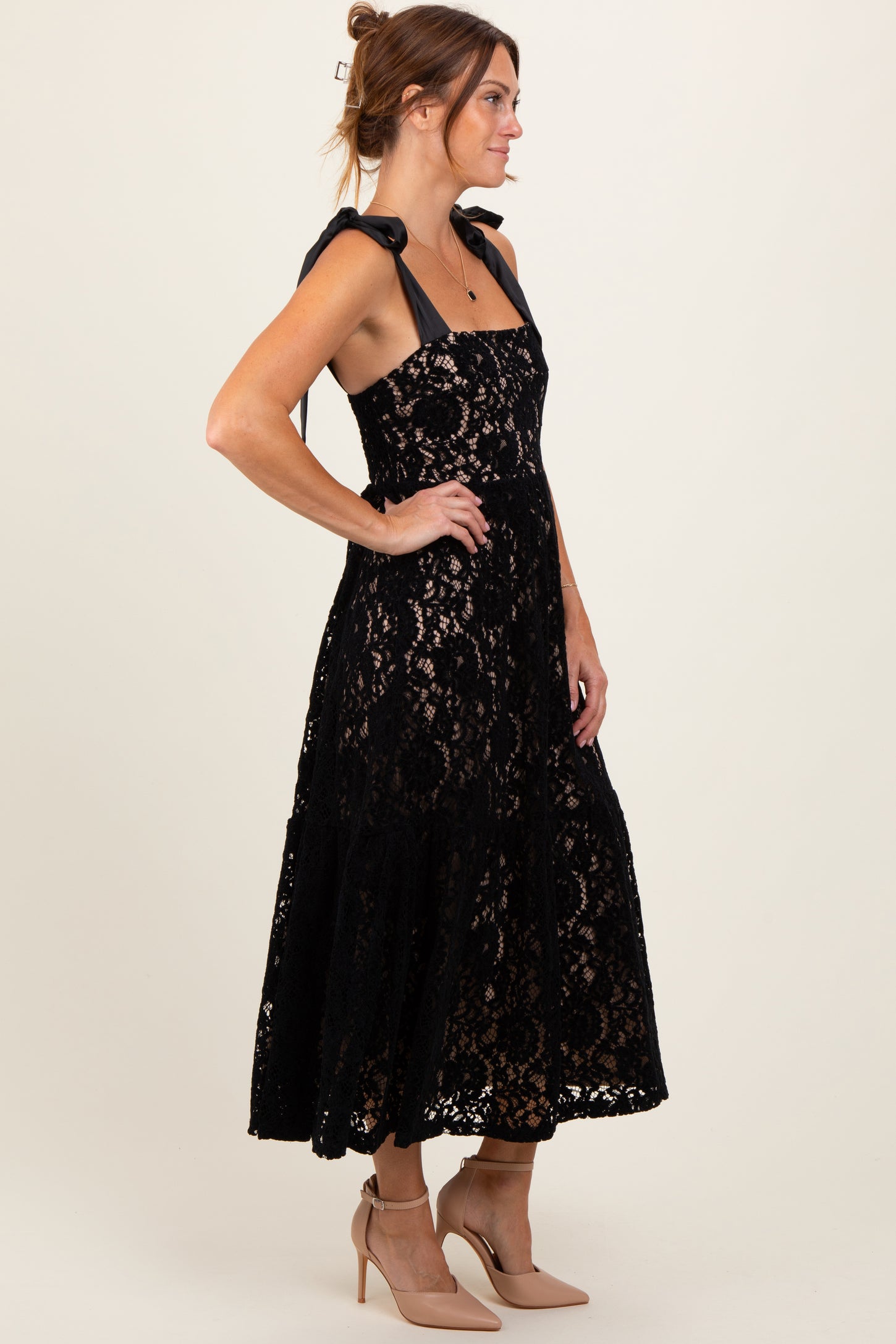 woman wearing a black lace midi dress with bow tie straps and nude heels, posing in profile