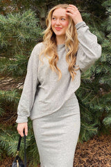 woman wearing a gray oversized sweater and a fitted gray midi skirt, standing outdoors near greenery
