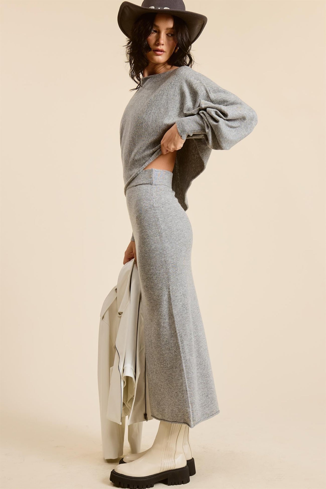woman wearing a gray oversized sweater and matching long skirt, paired with a hat, holding a jacket