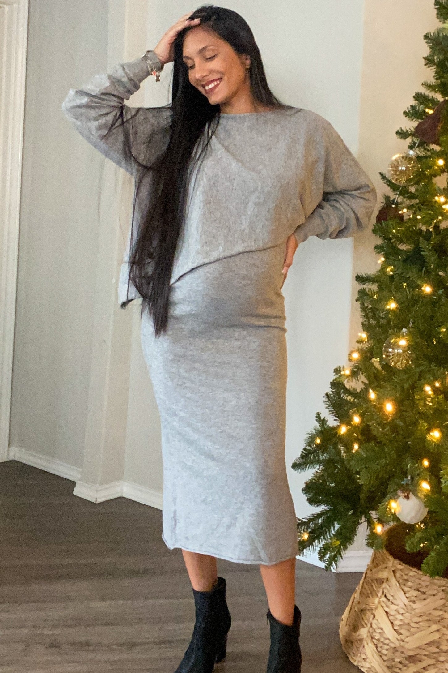 woman wearing a gray maternity dress with a cropped sweatshirt style and a visible baby bump, posing next to a Christmas tree