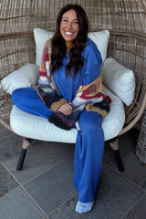 woman wearing a blue loungewear set with a multicolored chunky knit cardigan, sitting on a chair with a smile
