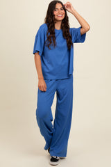 woman wearing a blue ribbed short sleeve top and matching wide-leg pants, smiling and posing with one hand raised