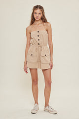 woman wearing a beige sleeveless romper with buttons and pocket details, styled with a cinched waist and casual sneakers