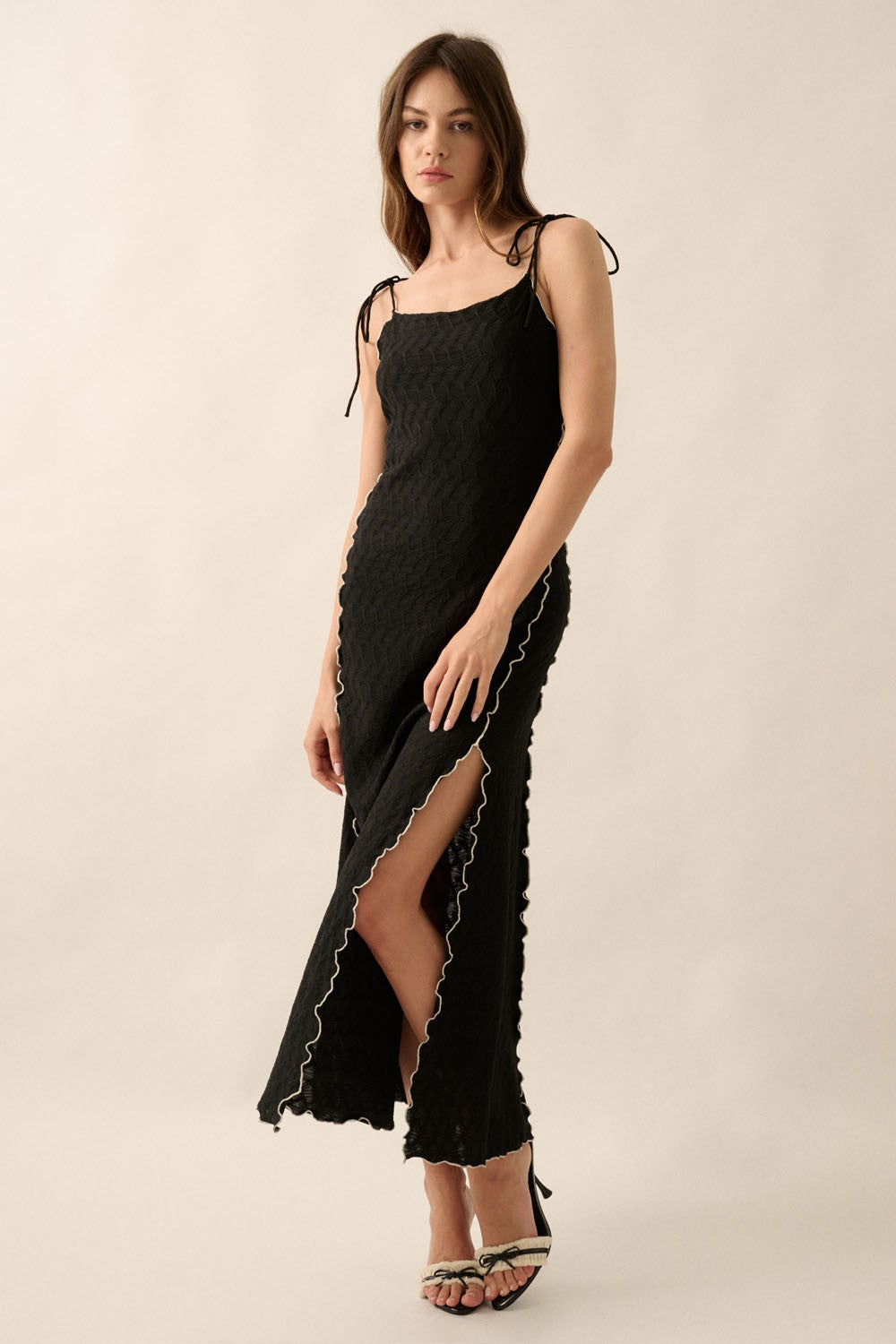 woman wearing a black maxi dress with tie shoulder straps and scalloped edges, featuring a thigh-high slit
