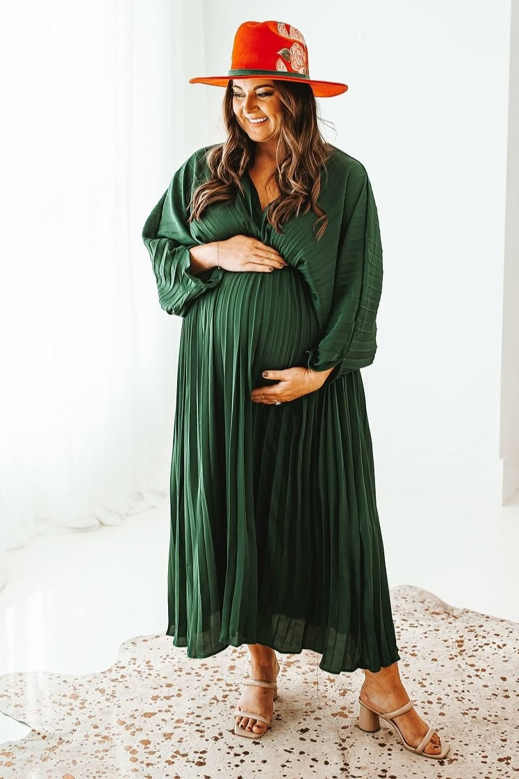 woman wearing a green pleated maternity dress and a red hat, holding her baby bump