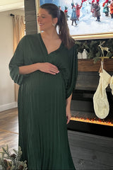 woman wearing a dark green pleated maternity gown, gently holding her baby bump while smiling