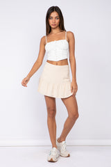 woman wearing a white crop top with buttons and a beige ruffled mini skirt, standing with one foot forward