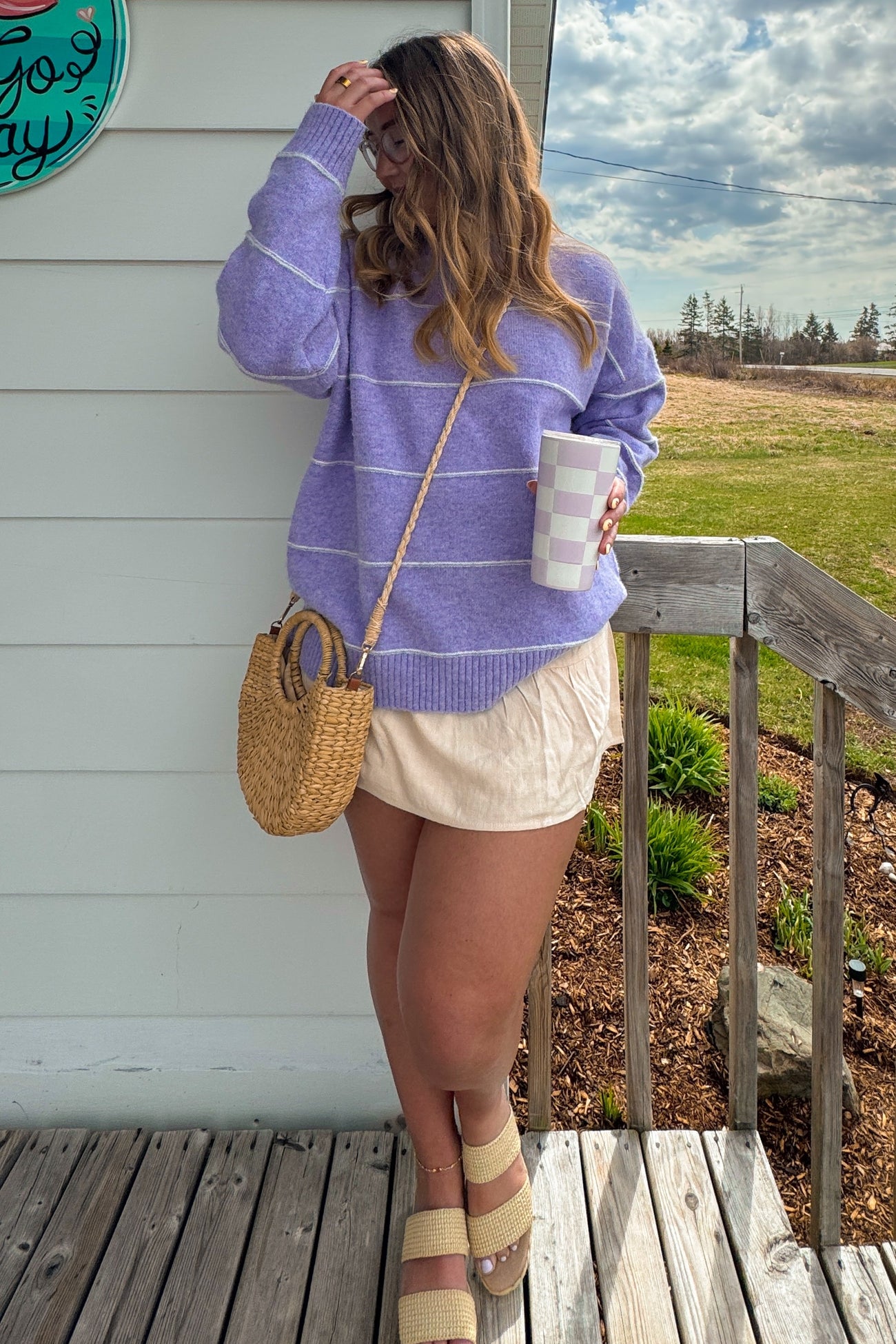 woman wearing a lavender striped sweater and beige shorts, holding a cup, standing on a porch with a straw bag and sandals