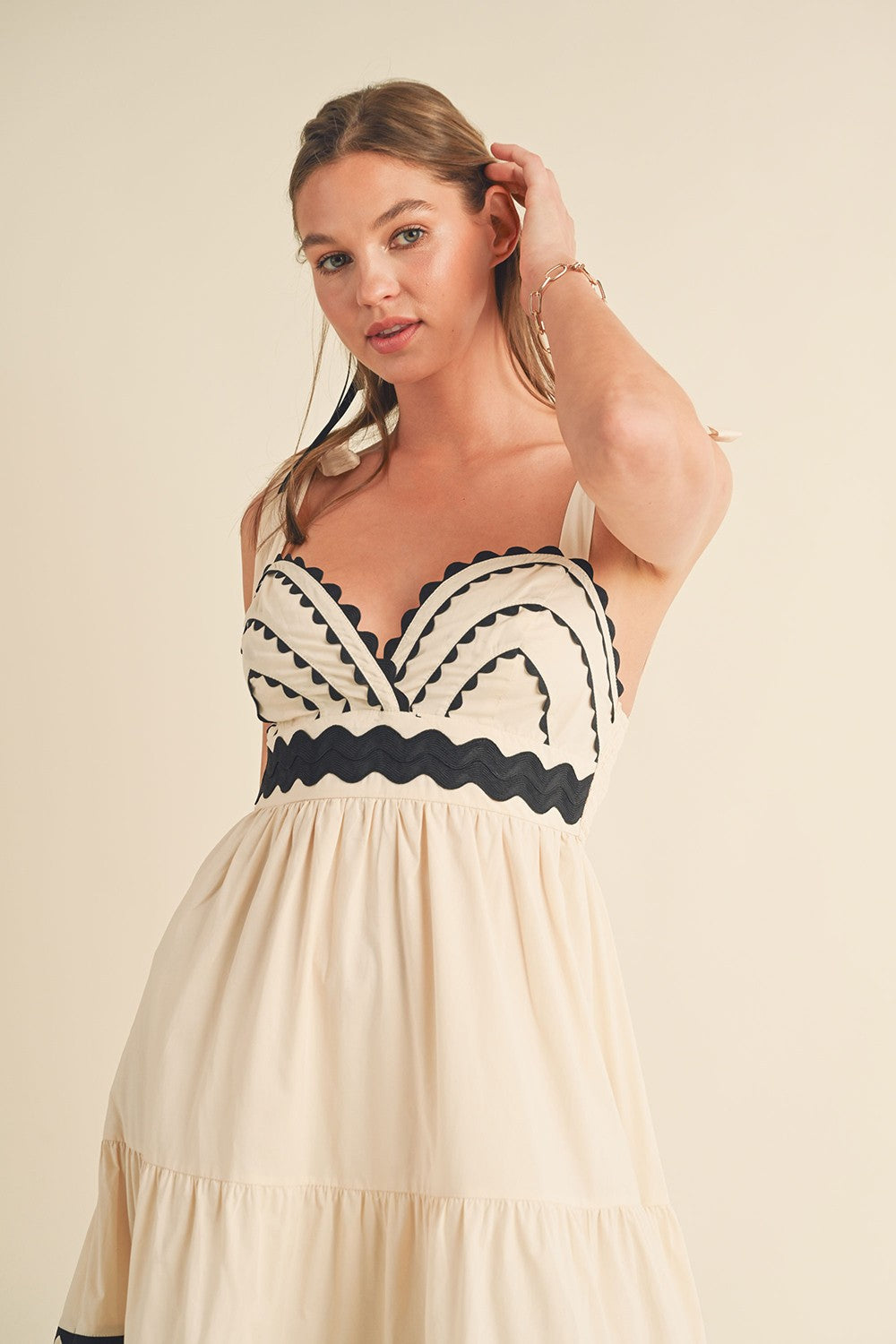 woman wearing a cream-colored sundress with black scalloped details and a fitted bodice, posing with one hand in her hair