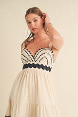 woman wearing a cream-colored sundress with black scalloped details and a fitted bodice, posing with one hand in her hair