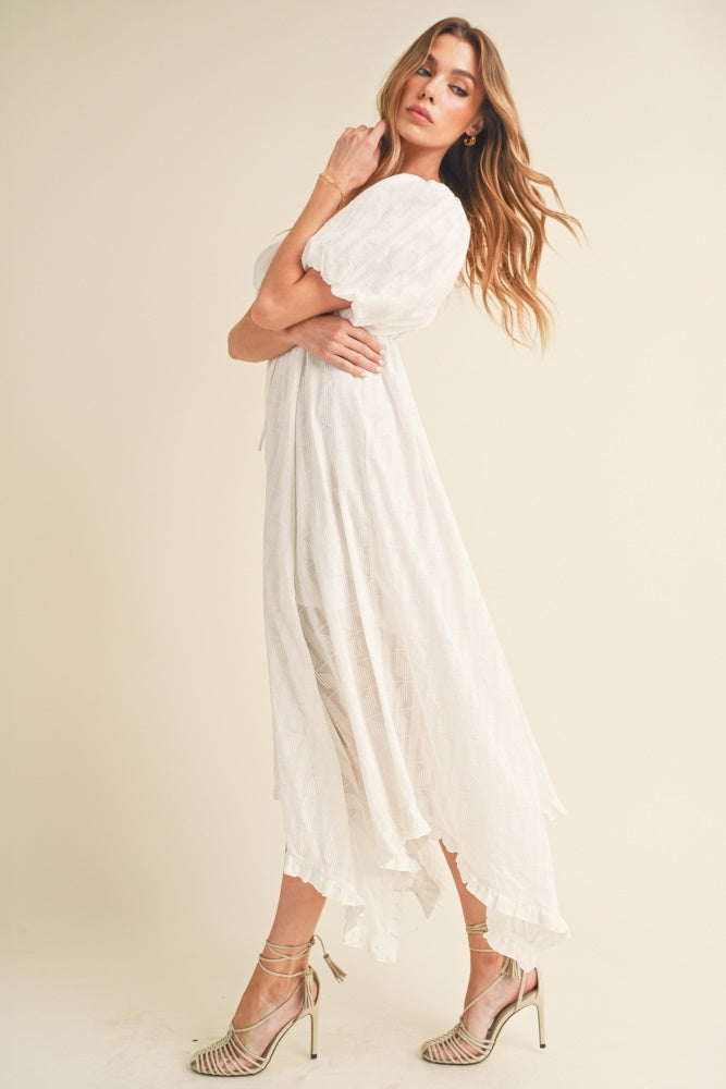 woman wearing a white flowy maxi dress with puff sleeves and high-low hem, paired with strappy heels
