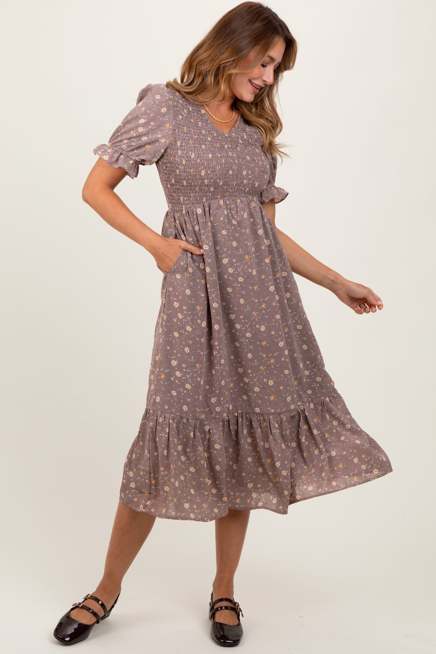 woman wearing a brown floral midi dress with puff sleeves and pockets, smiling while standing to the side