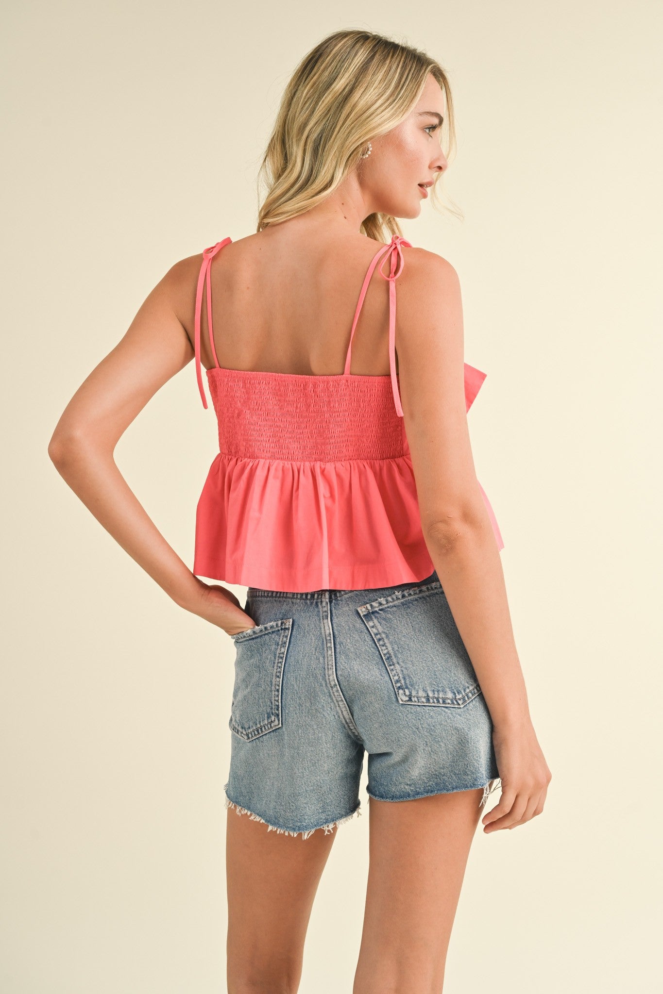 woman wearing a coral peplum top with adjustable straps and smocked back, paired with denim shorts