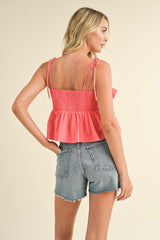 woman wearing a coral peplum top with adjustable straps and smocked back, paired with denim shorts