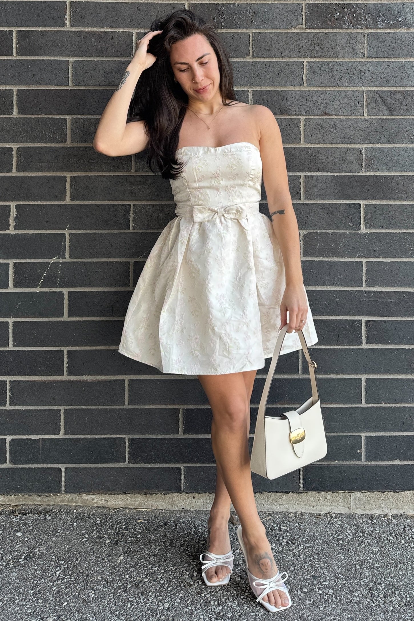 woman wearing a strapless cream-colored fit-and-flare dress with a bow detail, holding a white handbag and wearing strappy sandals
