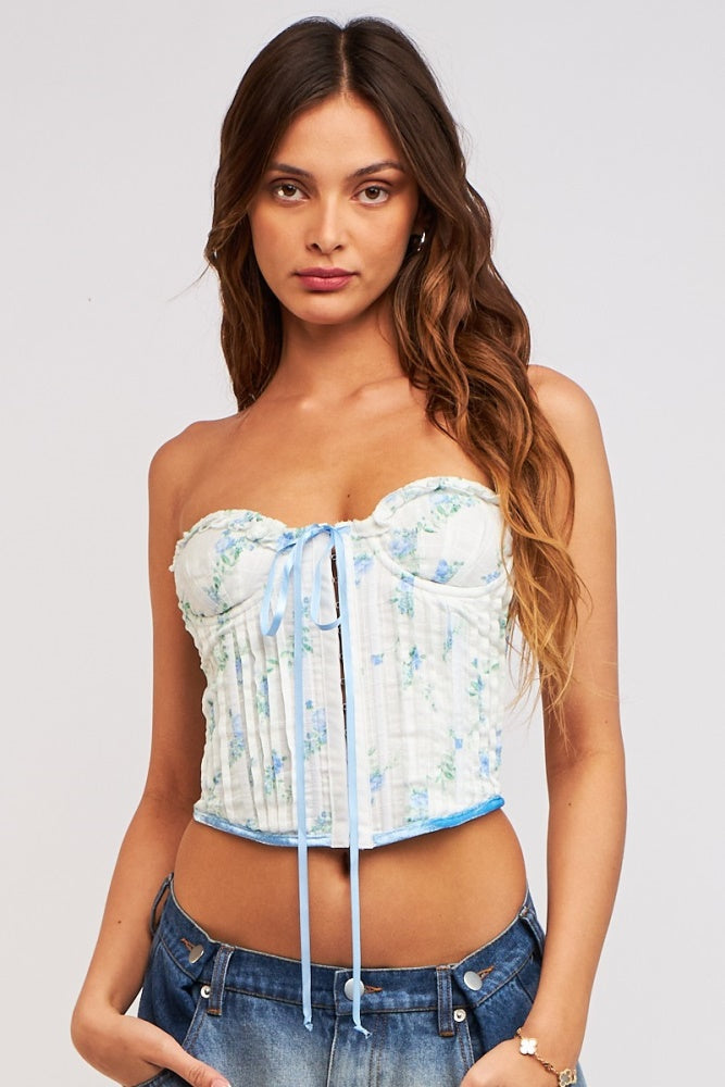 woman wearing a white floral corset top with blue accents and lace-up front, paired with denim jeans