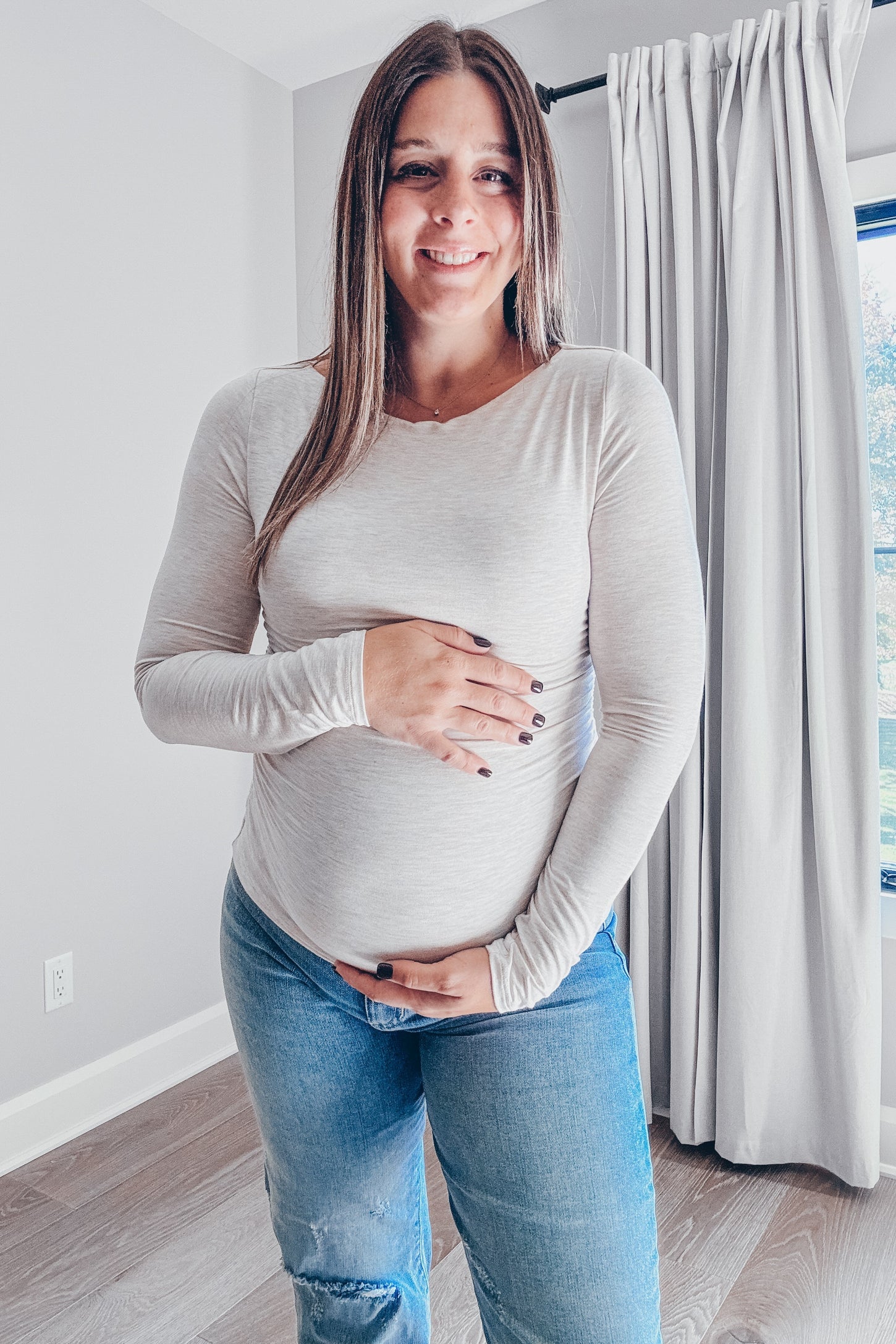 woman wearing a light beige long sleeve maternity top and holding her baby bump while standing in a bright room