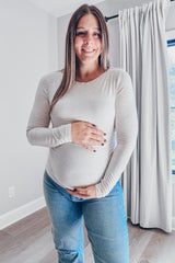 woman wearing a light beige long sleeve maternity top and holding her baby bump while standing in a bright room