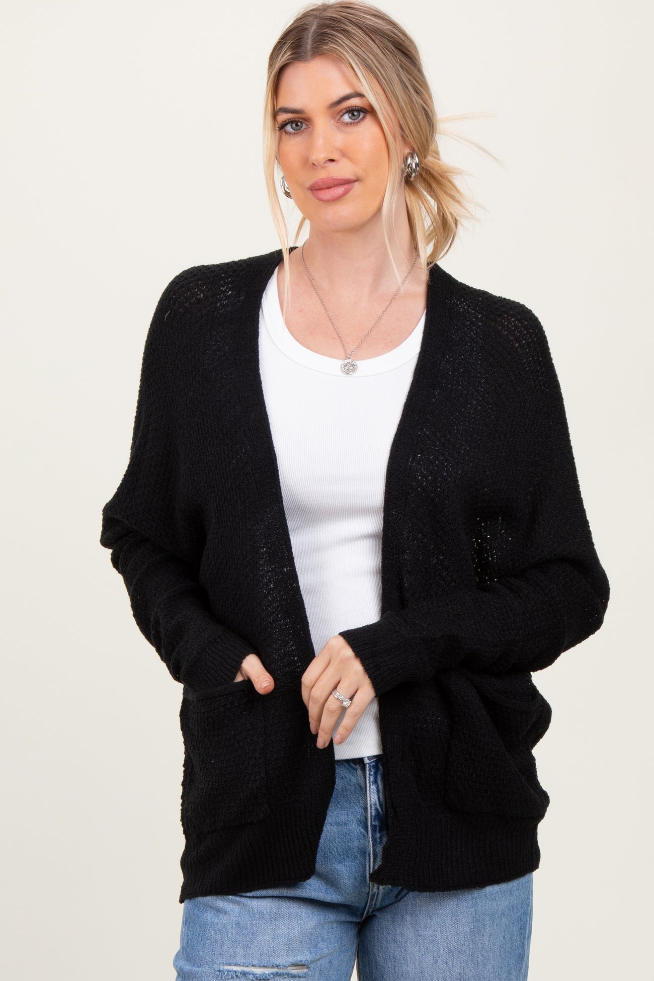 woman wearing a black knitted cardigan over a white top and blue jeans, with hands in pockets and a relaxed expression