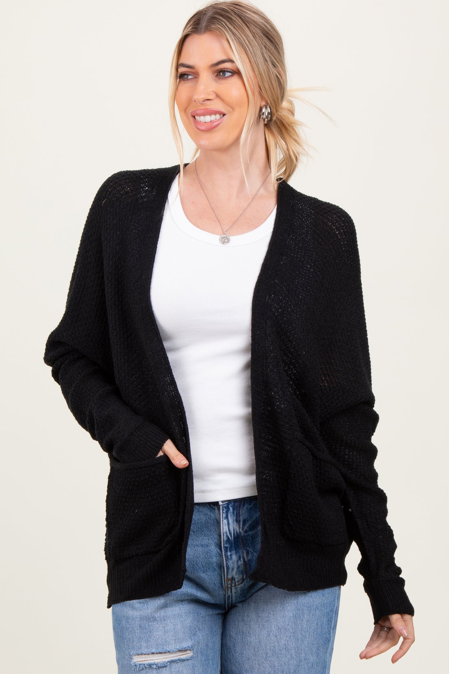 woman wearing a black open-knit cardigan over a white tank top and blue jeans, smiling with hands in pockets