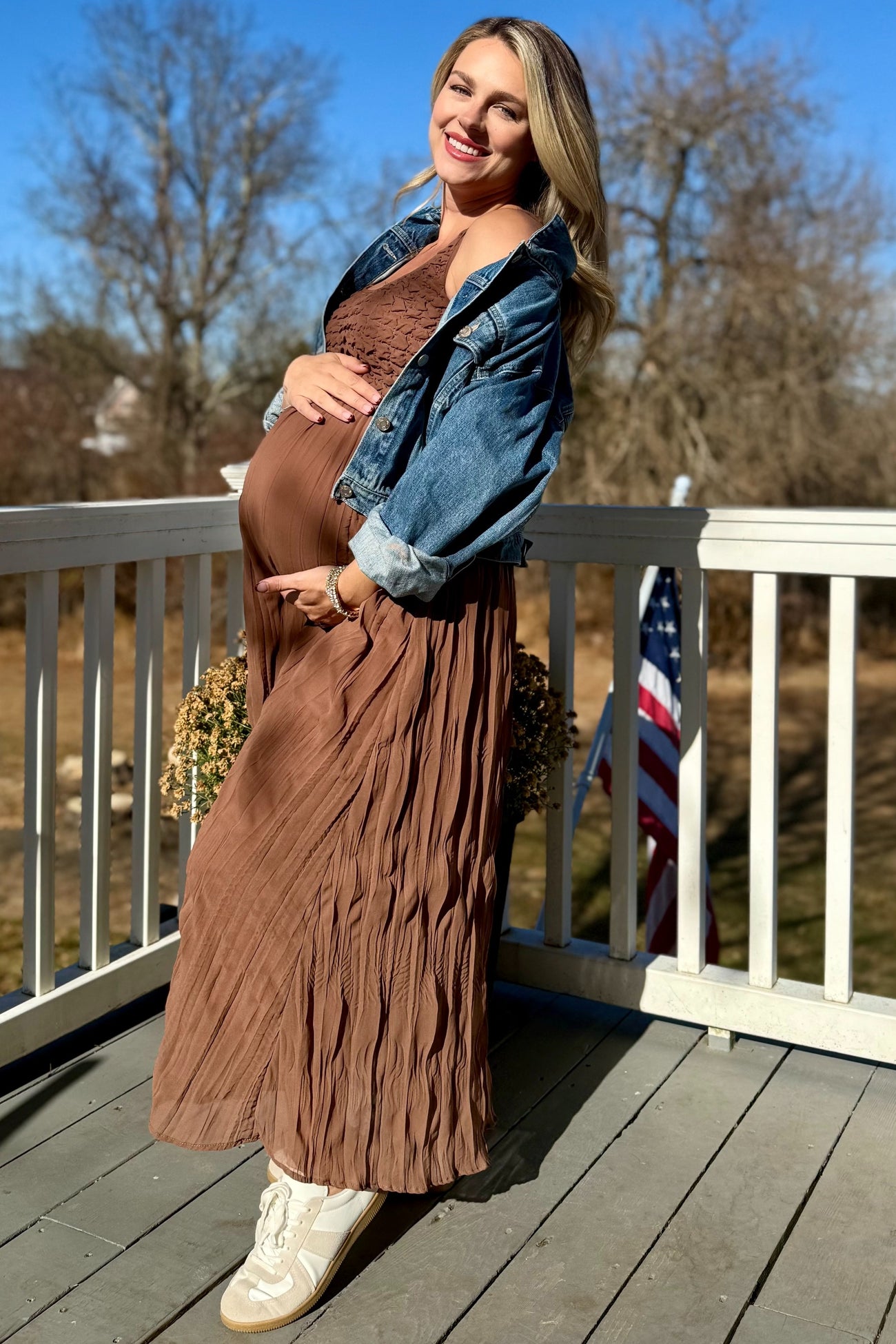 woman wearing a brown pleated maternity dress and a denim jacket, showing her baby bump while standing outdoors